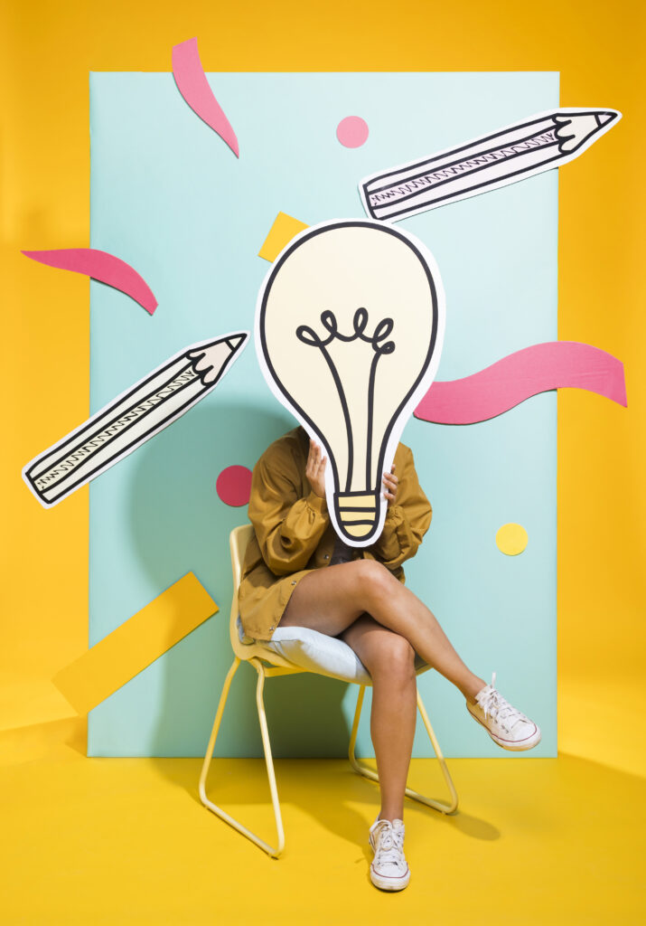 Das Bild zeigt ein junges Mädchen, dass auf einen Sessel sitzt und vor ihrem Gesicht hält sie ein gezeichnetes Bild einer Glühbirne.